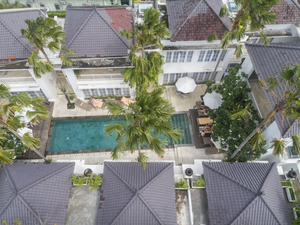 Aerial view of the swimming pool at The Colony Hotel Bali, featuring colonial-style architecture, surrounded by lush tropical plants and sun lounges