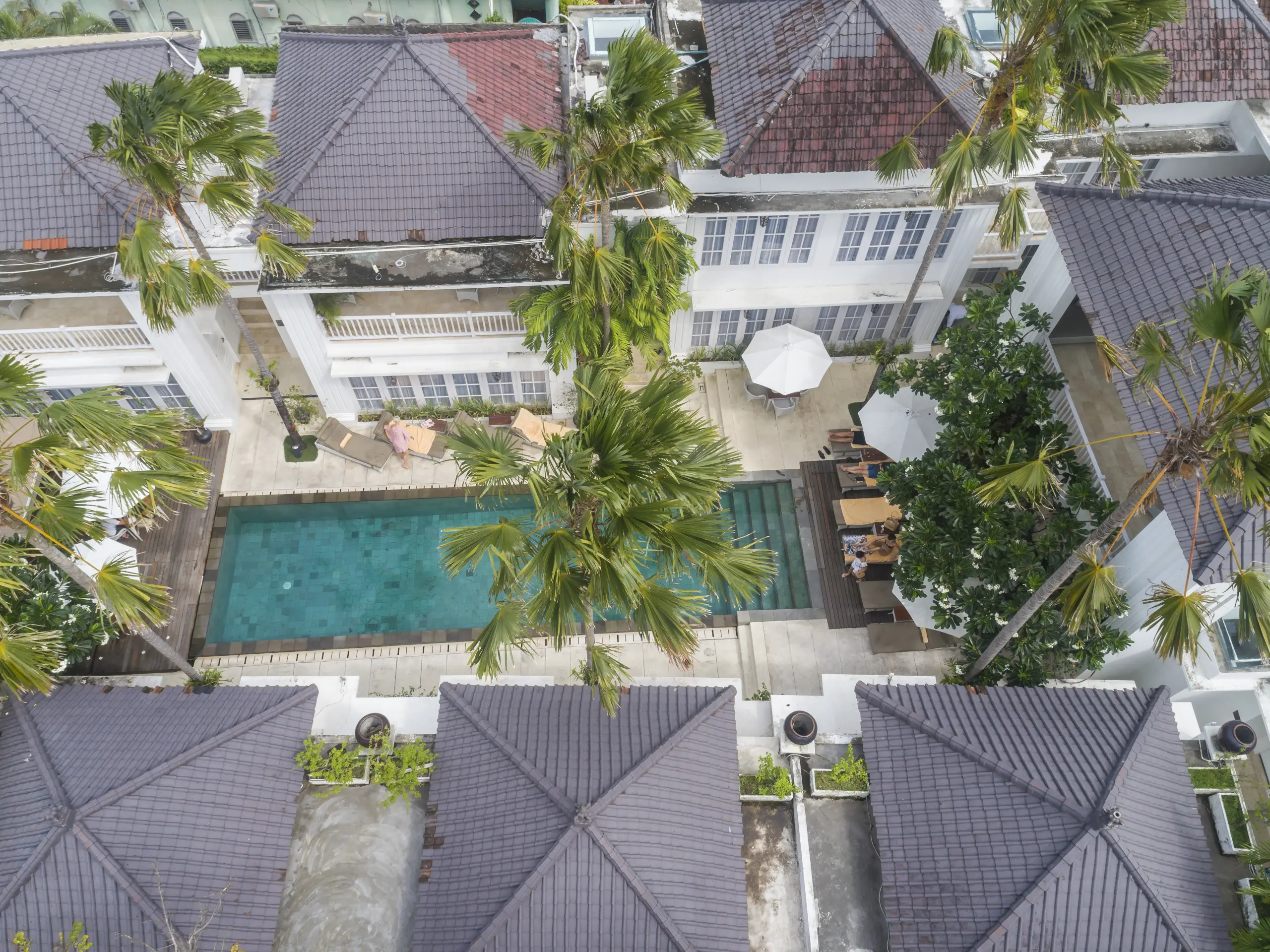 Aerial view of the swimming pool at The Colony Hotel Bali, featuring colonial-style architecture, surrounded by lush tropical plants and sun lounges