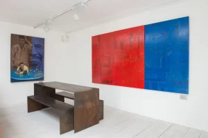 Minimalist white gallery room displaying bold red and blue abstract paintings alongside a rustic metal bench
