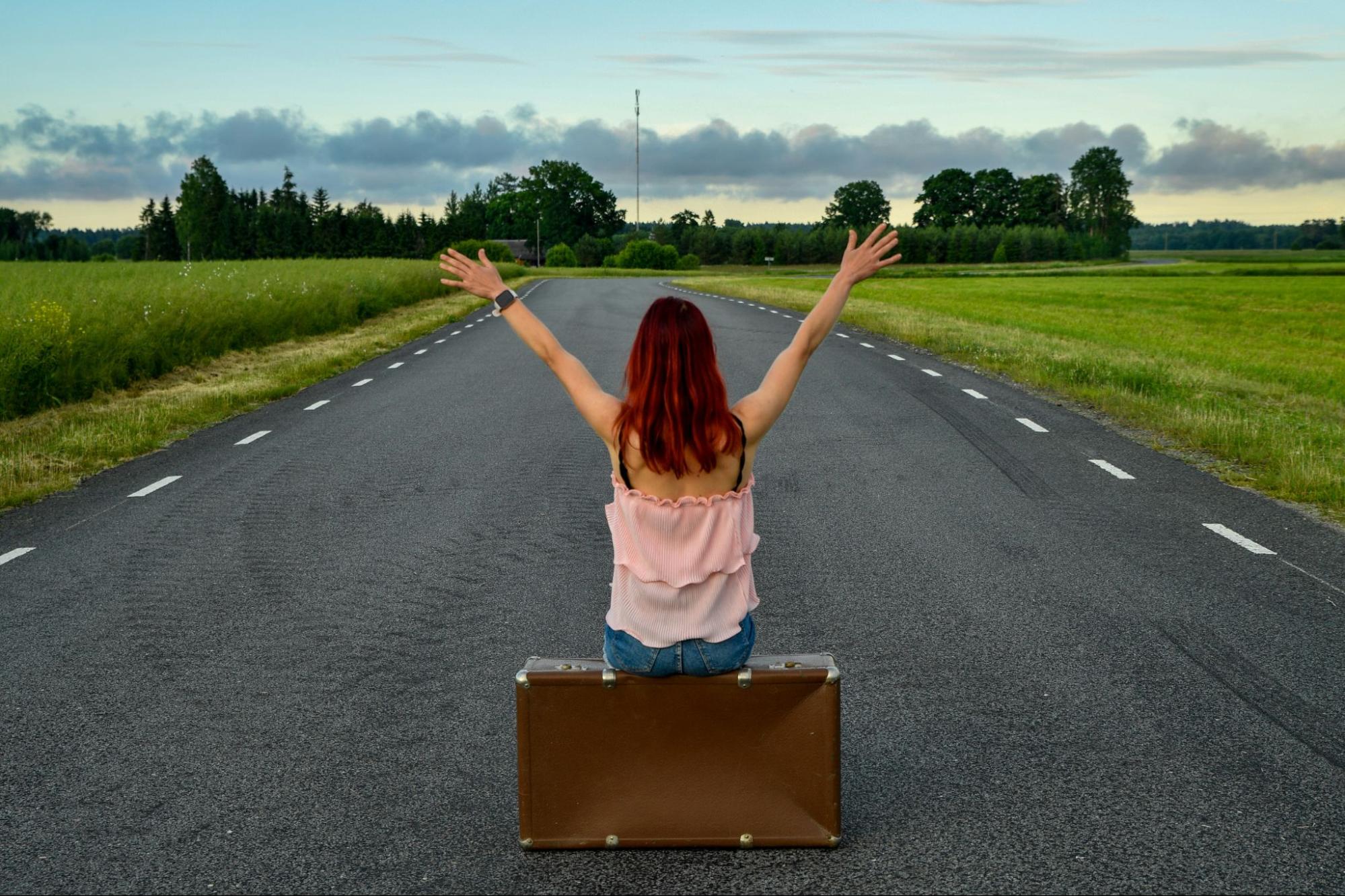 Woman with red hair sitting on suitcase with arms raised on empty scenic road at sunset