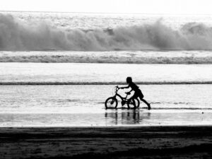 Black and white beach scene with child riding a bicycle along the shoreline
